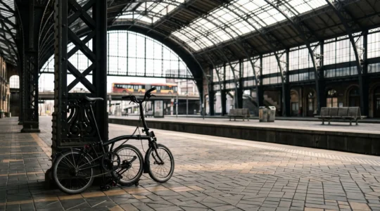 Vélo pliant, quai de gare belge et bus en arrière-plan symbolisant l'intermodalité pour réduire les frais de transport